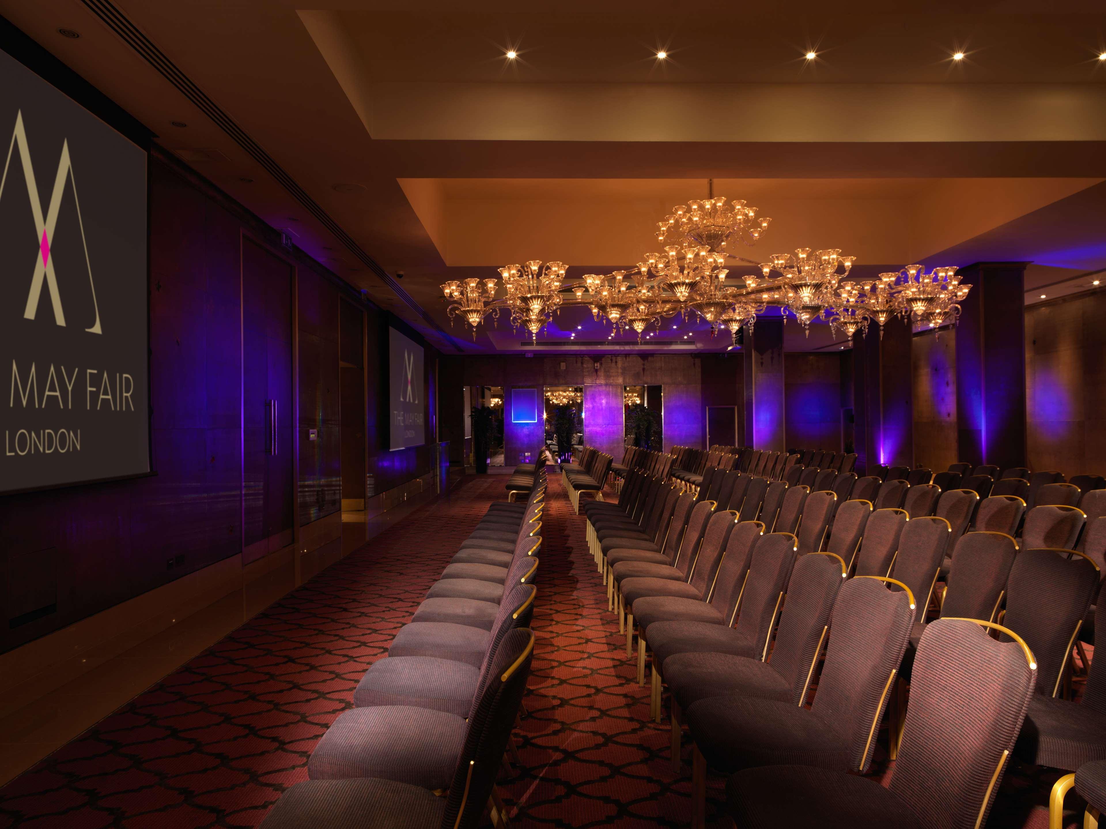 The May Fair, A Radisson Collection Hotel, Mayfair Londres Extérieur photo The image shows a conference or event space with rows of elegant, upholstered chairs facing a large screen. The room is adorned with chandeliers that provide ambient lighting, and there are purple lighting accents around the space. The branding for "