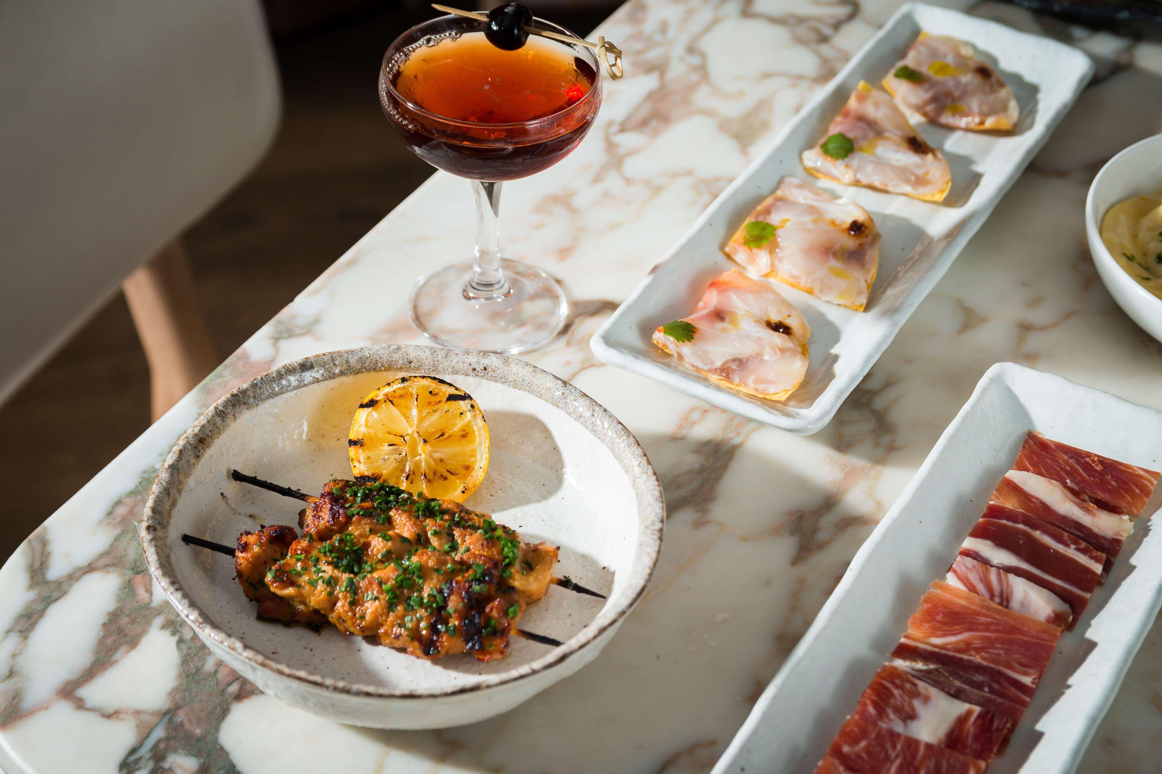 The May Fair, A Radisson Collection Hotel, Mayfair Londres Extérieur photo The photo features a beautifully arranged dining table with a variety of food and a drink. In the foreground, there is a dish containing grilled or cooked meat, garnished with herbs and served with a roasted lemon slice. Next to it, a cocktail drink 