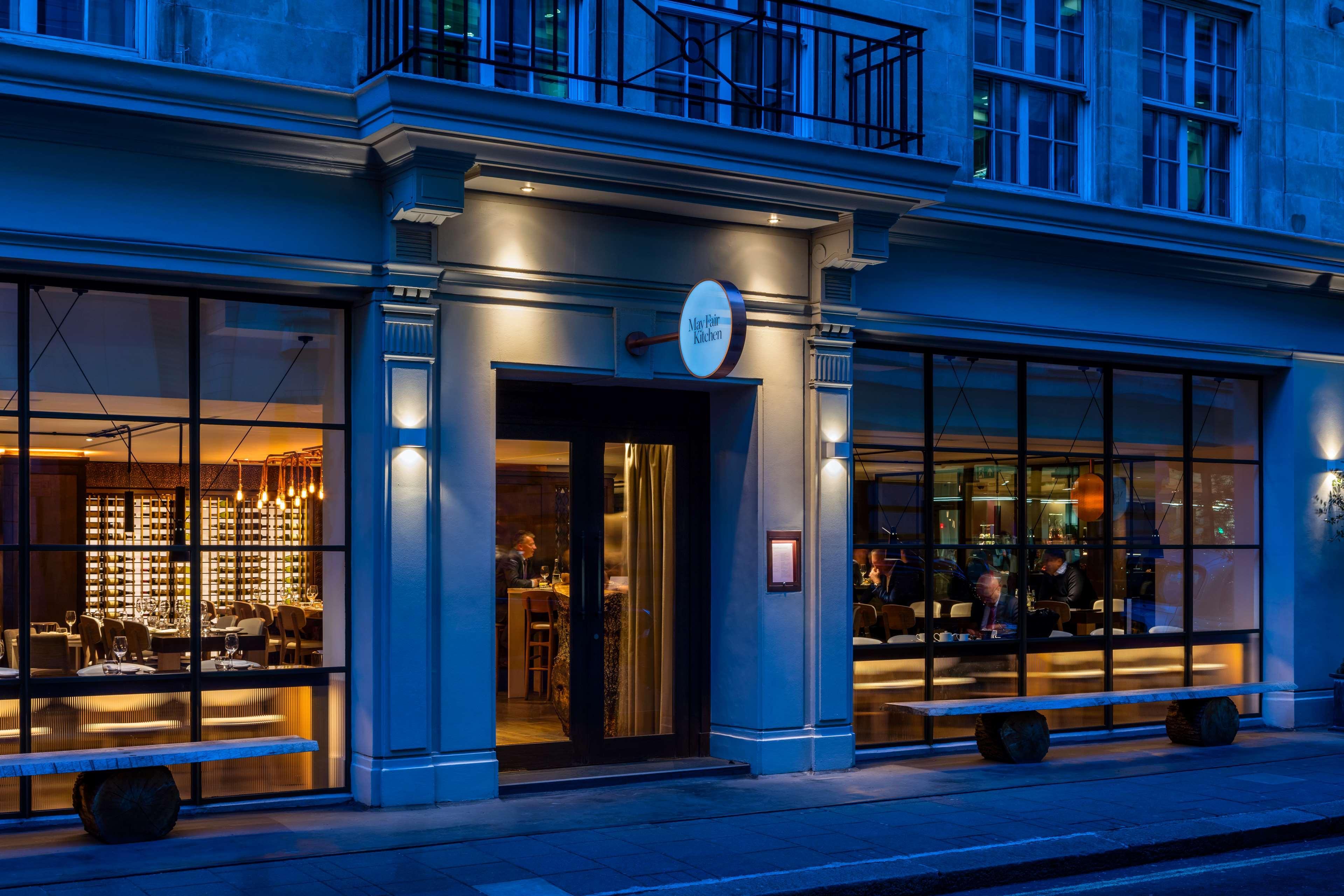The May Fair, A Radisson Collection Hotel, Mayfair Londres Extérieur photo The photo shows the exterior of a stylish restaurant. The entrance features a glass door and large windows, allowing a view inside. The restaurant is illuminated with soft lighting, creating a warm and inviting atmosphere against the evening backdrop
