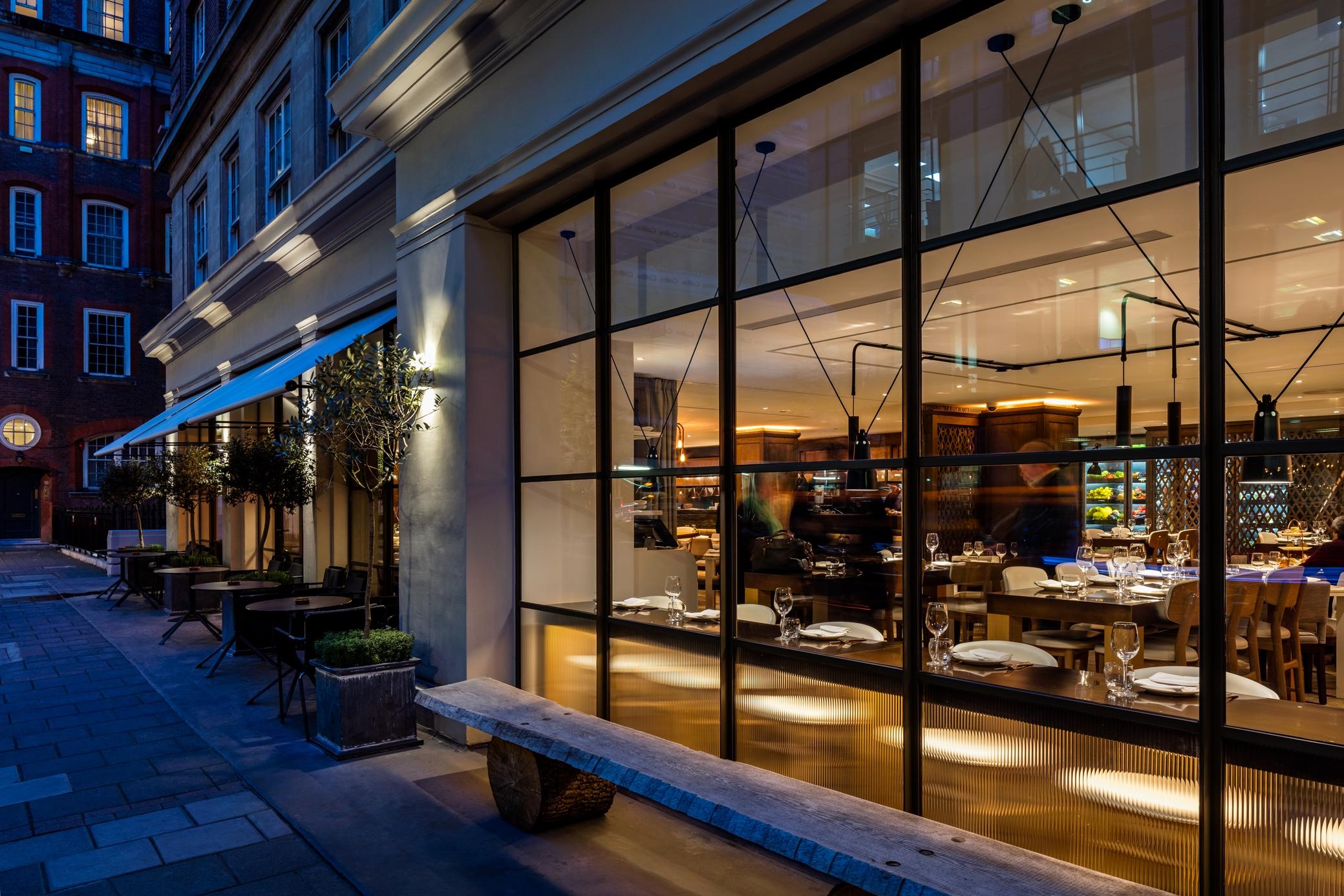 The May Fair, A Radisson Collection Hotel, Mayfair Londres Extérieur photo The photo shows the exterior of a restaurant viewed from the outside at night. It features large glass windows that allow a view of the interior, which is warmly lit. Inside, there are tables set for dining, indicating a welcoming atmosphere. Outside