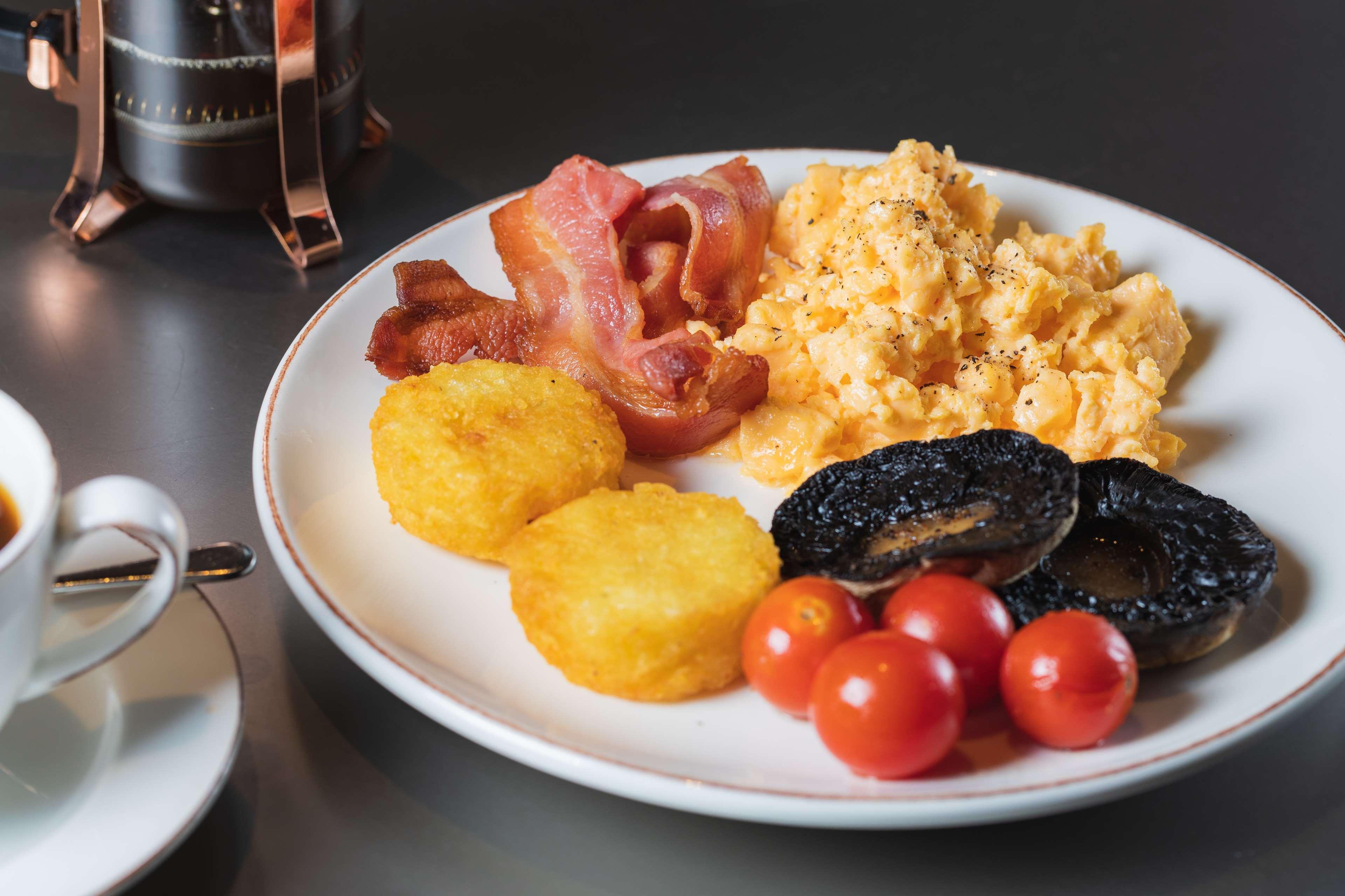 The May Fair, A Radisson Collection Hotel, Mayfair Londres Extérieur photo The photo shows a plate of breakfast food that includes scrambled eggs, crispy bacon strips, two golden hash browns, grilled tomatoes, and slices of black pudding. The dish appears to be served alongside a small cup, presumably for coffee or tea. The