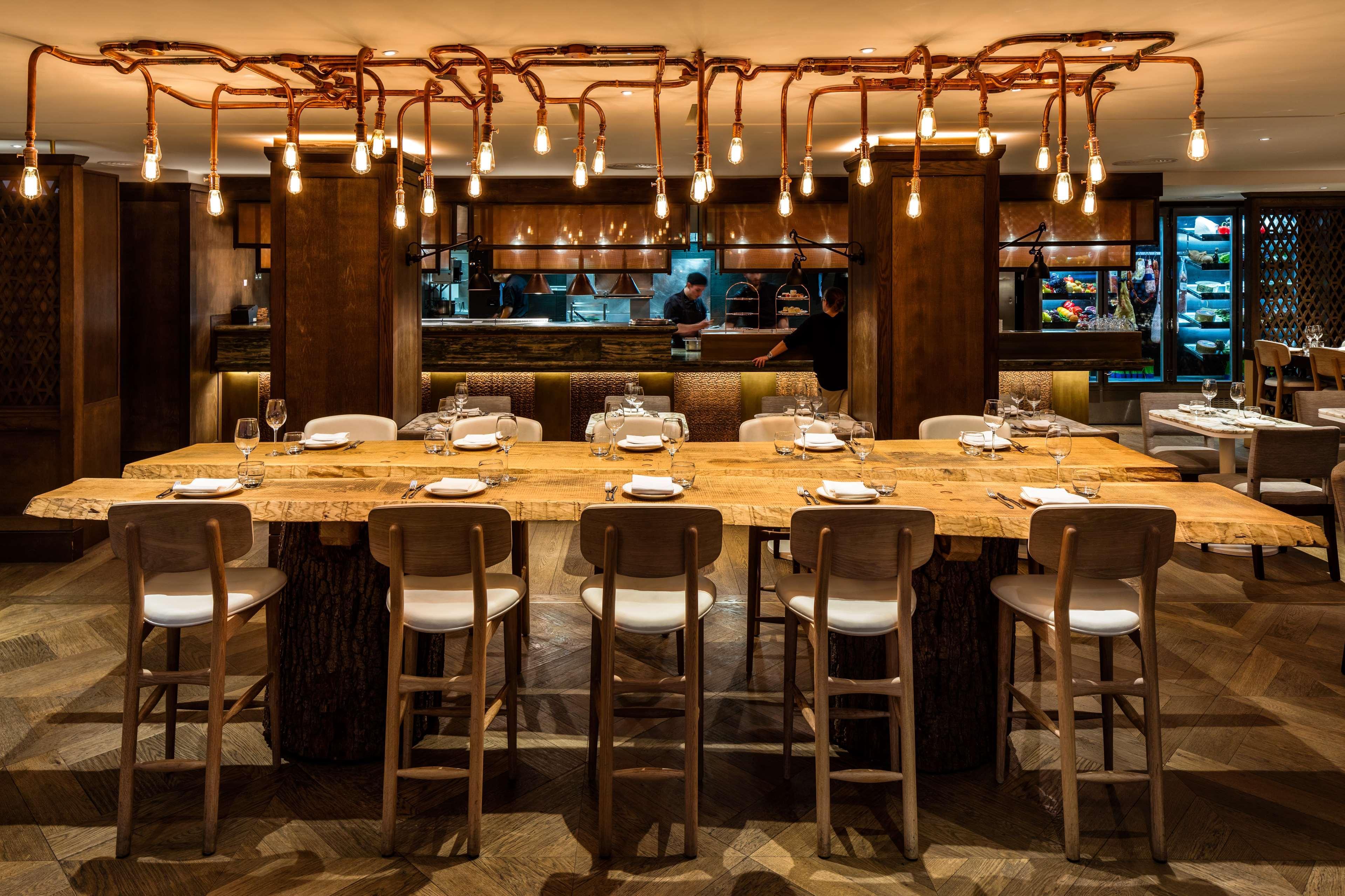 The May Fair, A Radisson Collection Hotel, Mayfair Londres Extérieur photo The photo shows a stylish restaurant interior. There is a large, rustic wooden dining table with several white chairs around it, suggesting a communal dining experience. Above the table, unique light fixtures made of twisted branches and hanging bulb