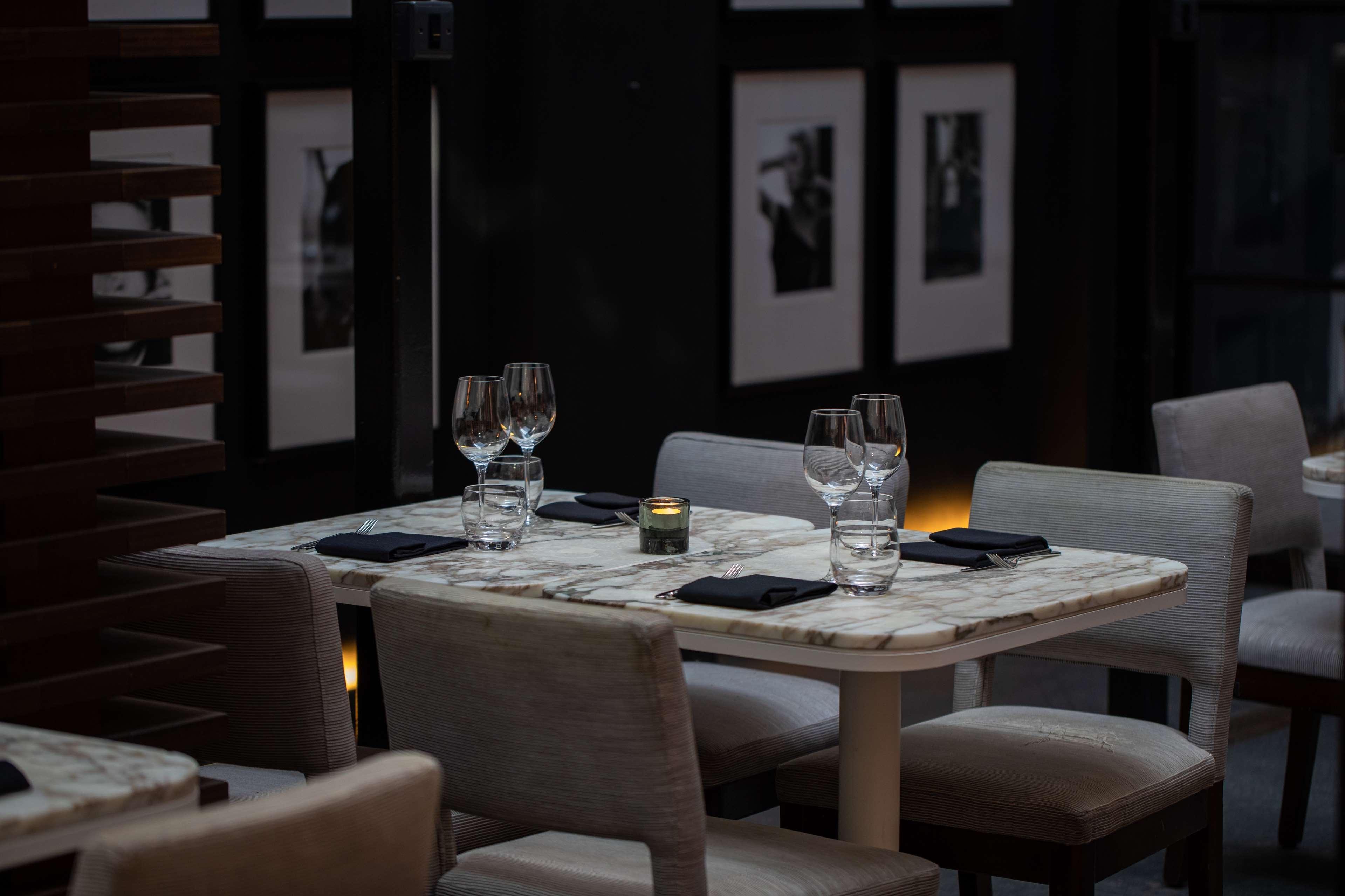 The May Fair, A Radisson Collection Hotel, Mayfair Londres Extérieur photo The photo shows a dining area with a stylish and elegant atmosphere. There are two tables set for dining, featuring marble tops and light-colored chairs. Each table has wine glasses and a small candle holder, creating a warm ambiance. In the backgrou