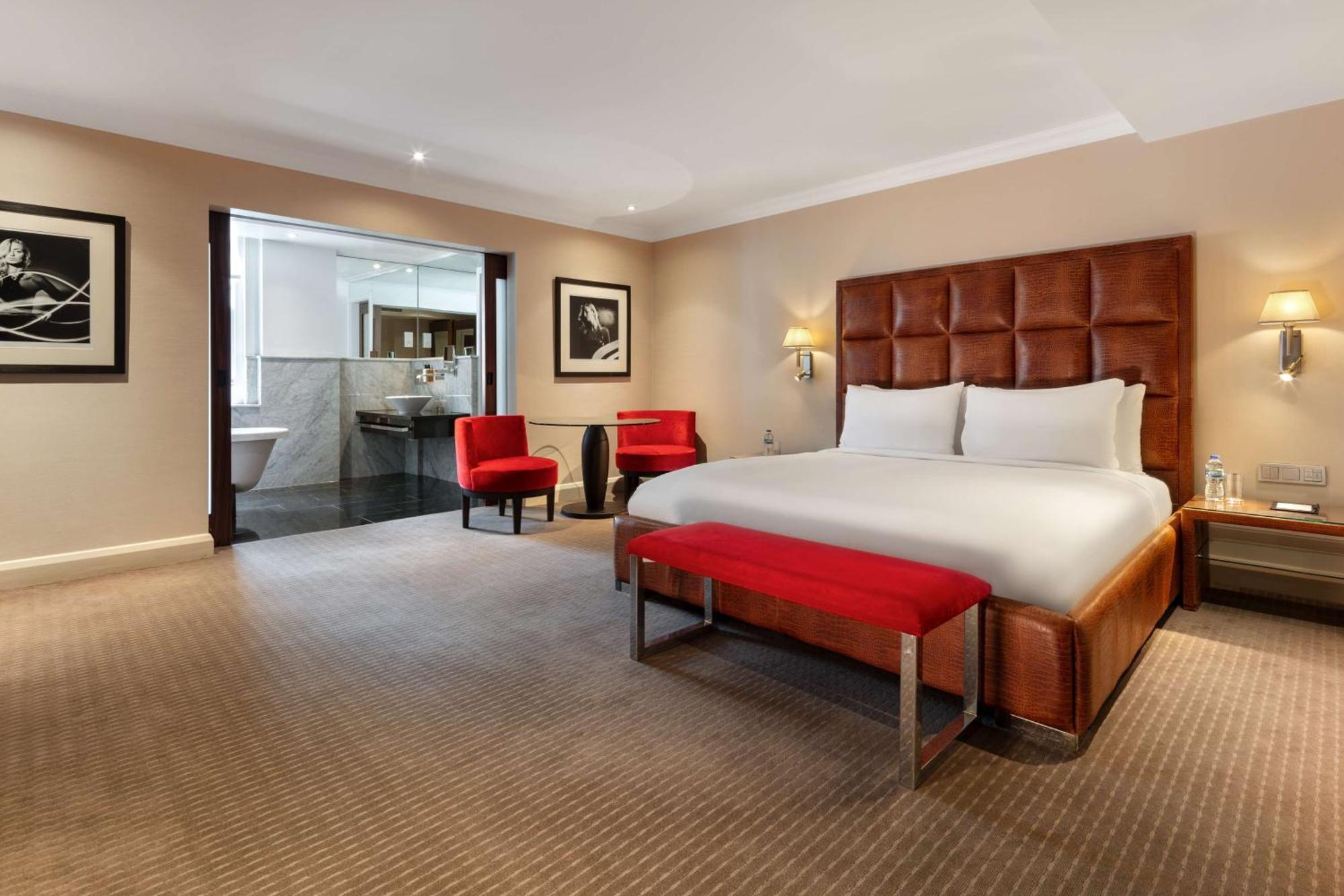 The May Fair, A Radisson Collection Hotel, Mayfair Londres Extérieur photo The photo shows a modern hotel room featuring a large, comfortable king-size bed with a beige and brown color scheme. There are two red accent chairs positioned near the bed, adding a pop of color. A small red bench is placed at the foot of the bed. 