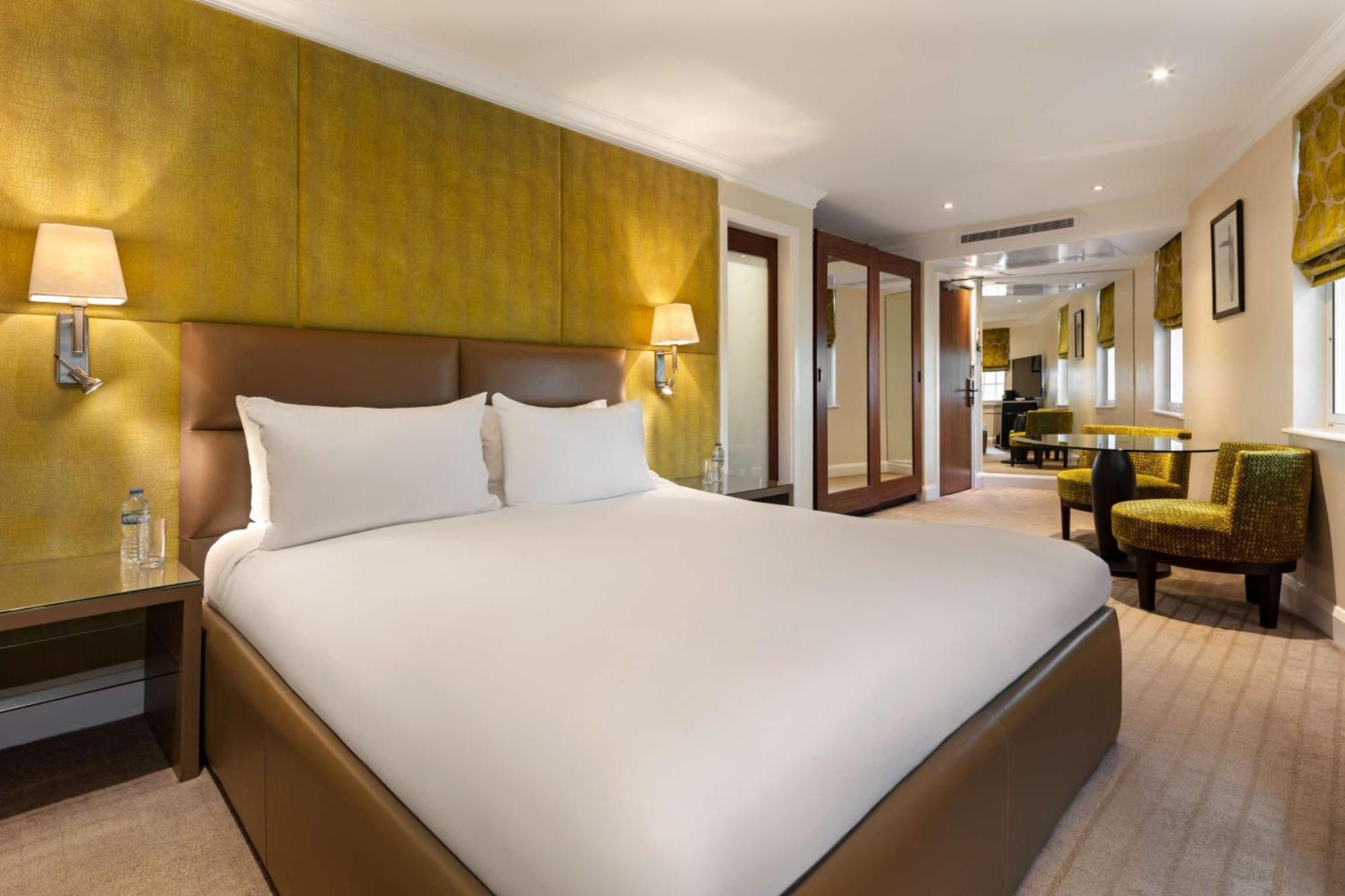 The May Fair, A Radisson Collection Hotel, Mayfair Londres Extérieur photo The photo shows a modern hotel room featuring a large bed with a neatly made white duvet and pillows. The walls are accented with a soft yellow color, enhancing the inviting atmosphere. On either side of the bed, there are bedside tables with lamps. 