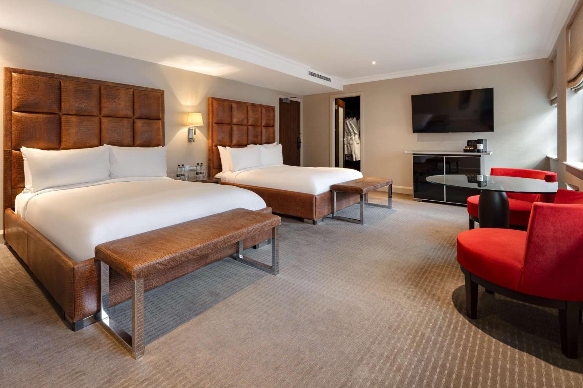 The May Fair, A Radisson Collection Hotel, Mayfair Londres Extérieur photo The photo shows a modern hotel room with two beds. Each bed is made with white linens and has a large, upholstered headboard. There is a bench at the foot of each bed. To the right, there are two red chairs around a small glass table. A flat-screen T