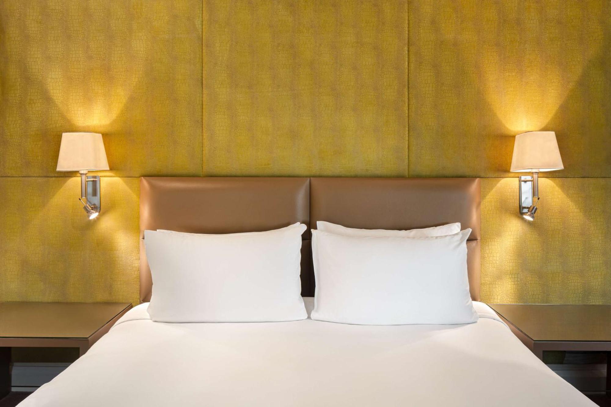 The May Fair, A Radisson Collection Hotel, Mayfair Londres Extérieur photo The photo shows a cozy bedroom setting featuring a large bed with two white pillows. The headboard has a smooth, light brown leather material, and there are two wall-mounted lamps on either side of the bed, illuminating a textured yellow wall behind 