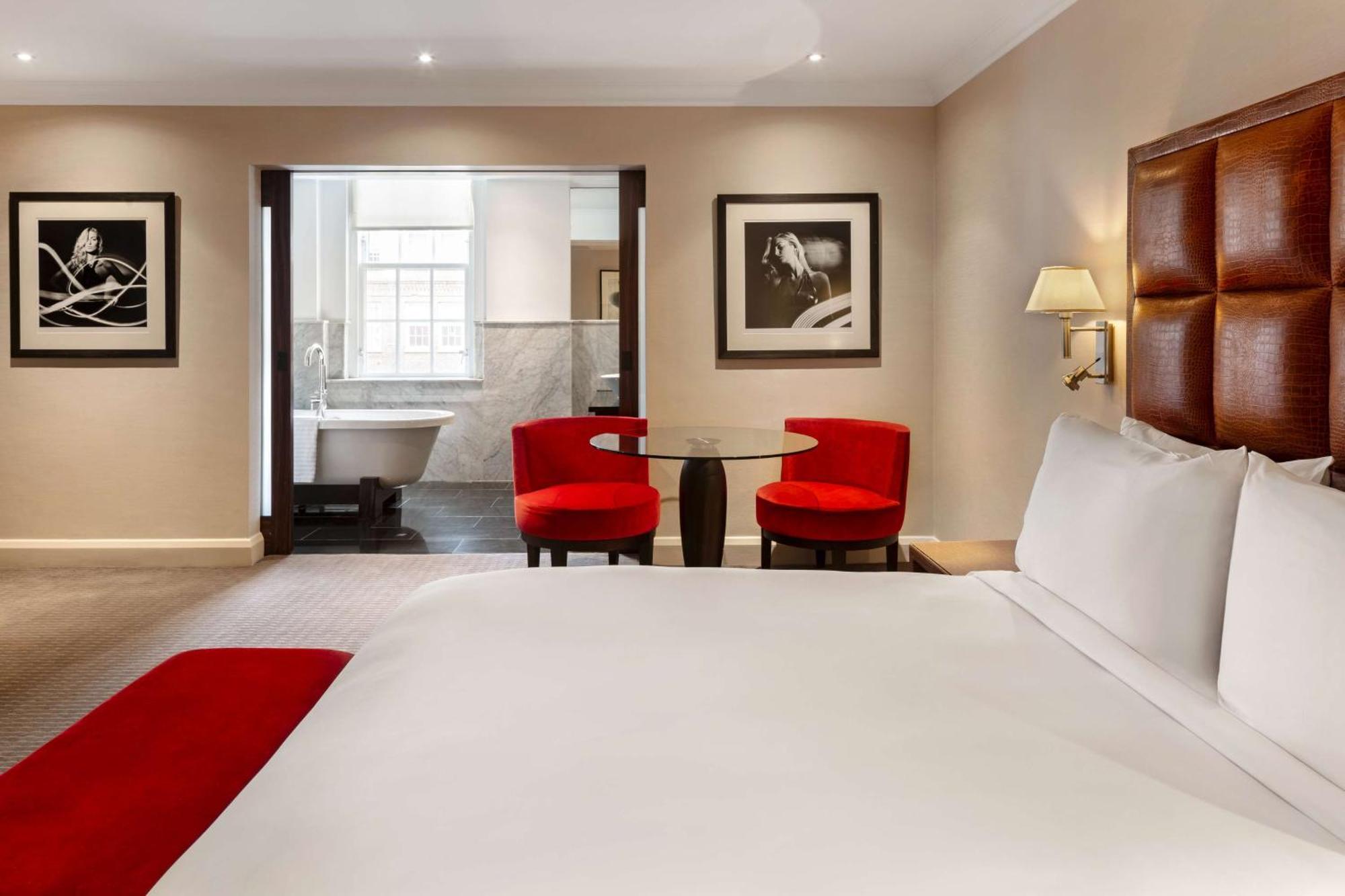 The May Fair, A Radisson Collection Hotel, Mayfair Londres Extérieur photo The photo shows a hotel room featuring a large bed with white linens and a red accent throw. In the background, there are two modern red chairs around a small glass table. The wall displays framed black and white photographs. To the side, there is an