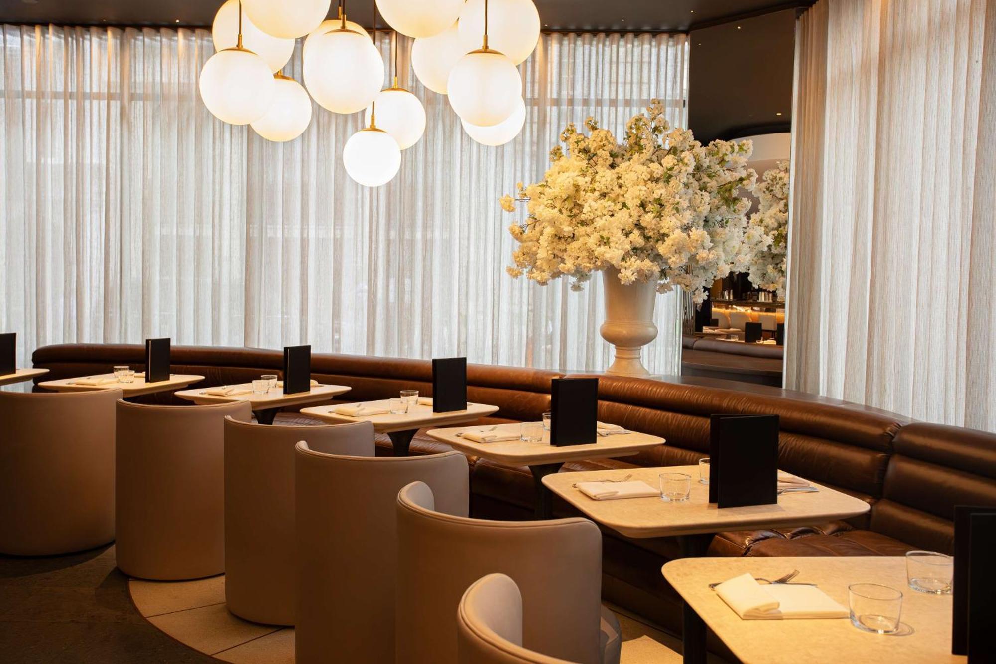 The May Fair, A Radisson Collection Hotel, Mayfair Londres Extérieur photo The photo shows a modern restaurant interior. It features stylish seating arrangements with curved booths and elegant tables. The lighting is soft, provided by large, round pendant lights hanging from the ceiling. A large floral arrangement in a whit