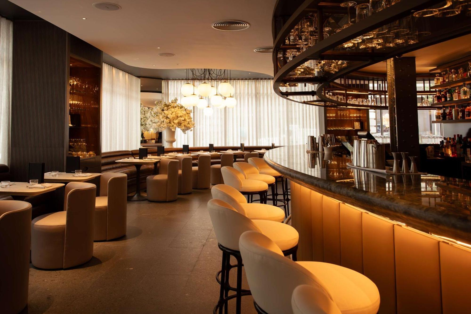 The May Fair, A Radisson Collection Hotel, Mayfair Londres Extérieur photo The photo shows the interior of a sleek and modern bar or restaurant. There is a curved bar counter with several stylish high stools in front of it. The seating area includes plush, rounded chairs arranged around circular tables. The design features 